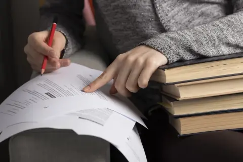 Ręka z ołówkiem wskazuje na dokumenty, stos książek obok. Czekanie na wyniki matur bywa stresujące.