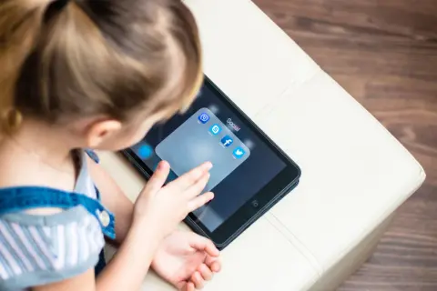 Dziecko dotyka ekranu tabletu z ikonami aplikacji społecznościowych, w tym Facebooka. Od ilu lat można mieć Facebooka?