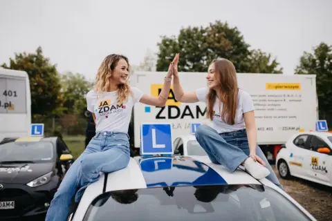 Dwie uśmiechnięte dziewczyny w koszulkach "Ja Zdam Szkoła Jazdy" świętują sukces, siedząc na dachu samochodu. To znak, że zdały egzamin klasyfikacyjny!