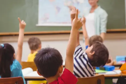 Uczniowie z podniesionymi rękami, gotowi do odpowiedzi. Nauczycielka ocenia ich zaangażowanie, co wpłynie na oceny z zachowania.