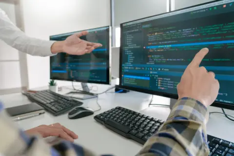 Dwóch programistów analizuje kod na monitorach. Jeden wskazuje palcem na ekran, drugi gestykuluje.