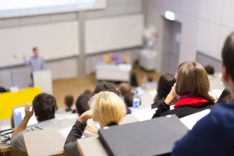 Nauczyciel akademicki prowadzi wykład w auli, studenci słuchają, niektórzy notują.