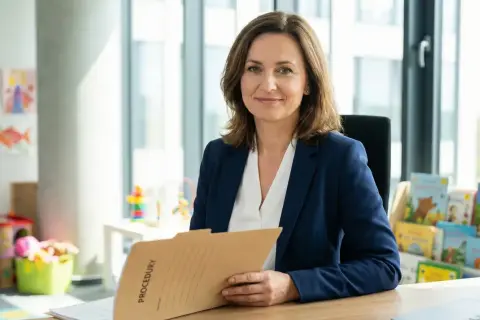 Kobieta w granatowej marynarce trzyma teczkę z napisem "PROCEDURY". W tle przedszkolne rysunki i książki. To przykładowy opis oceny pracy nauczyciela przedszkola.