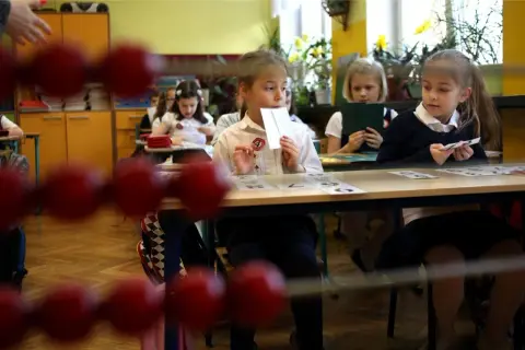Dzieci siedzą w ławkach, ucząc się, kiedy idą do szkoły. Jedna dziewczynka trzyma kartę z cyfrą.