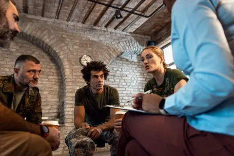 Grupa osób, w tym weterani, uczestniczy w sesji terapeutycznej. Rozmowa skupia się na wsparciu i procesie resocjalizacji.