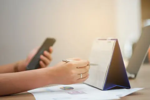 Ręka z długopisem zaznacza datę w kalendarzu, sprawdzając, czy 1 listopada jest dniem wolnym od pracy. Obok leży telefon.