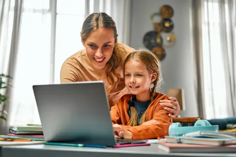 Uśmiechnięta mama pomaga córce w nauce przy laptopie. To przykład troskliwej opieki na dziecko 2 dni w tygodniu, gdy rodzice pracują.