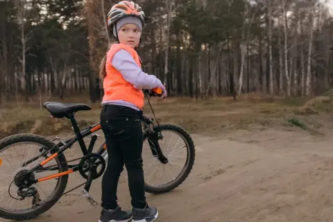 Dziewczynka w kasku i kamizelce przygotowuje się do egzaminu na kartę rowerową, stojąc obok swojego roweru na leśnej ścieżce.