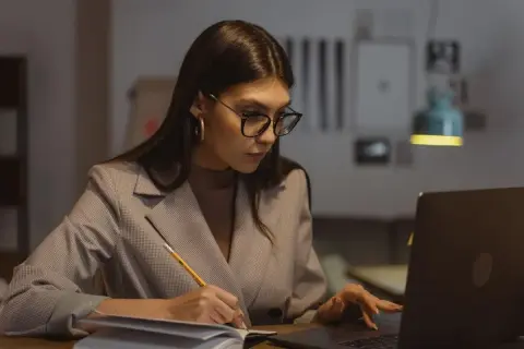 Kobieta w okularach pracuje przy laptopie, notując w zeszycie. Zastanawia się nad limitem nadgodzin w roku, by zachować równowagę.