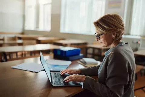 Kobieta w okularach pracuje na laptopie w pustej sali lekcyjnej. Może szukać informacji o studiach podyplomowych online bezpłatnych.
