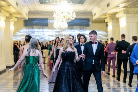 Młoda para idzie na studniówkę. Dziewczyna w czarnej, błyszczącej sukni z piórami, chłopak w eleganckim garniturze. W tle inni uczniowie w balowych strojach.