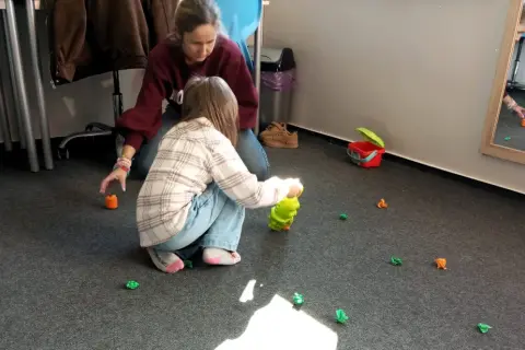 Dzieci bawią się na dywanie, zbierając małe, kolorowe figurki. W tle widać dorosłą osobę. To świetna zabawa z elementami wwr.