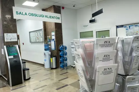 W sali obsługi klienta ZUS, obok dystrybutora wody, stoją stojaki z formularzami, m.in. ZUA, ZZA, ZCNA, RCA, ZUS RSA.
