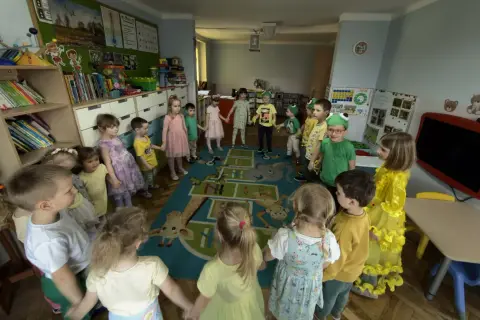 Dzieci trzymają się za ręce w kręgu na dywanie z zabawnymi zwierzątkami. To radosny dzień w **zespole szkolno przedszkolnym**.