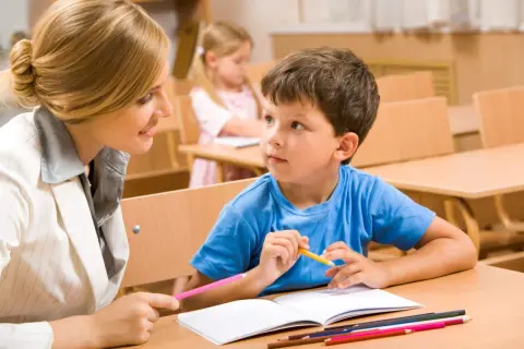 Nauczycielka wspiera chłopca z **specjalnymi potrzebami edukacyjnymi** w nauce. W tle widać inne dzieci.