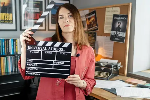 Kobieta trzyma klaps filmowy, gotowa do rozpoczęcia zajęć. W tle plakaty filmowe i maszyna do pisania, tworząc atmosferę pracy nad scenariuszem.