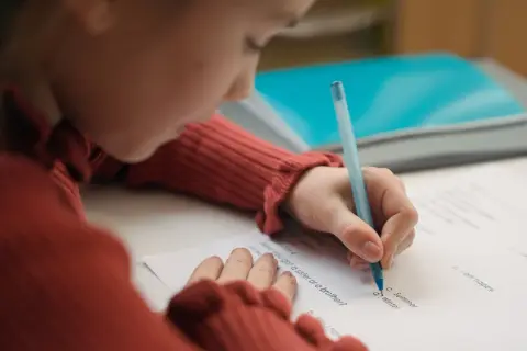 Dziecko w czerwonym swetrze pisze długopisem po karcie pracy, starając się o dopuszczający ocena.