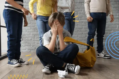 Chłopiec siedzi na podłodze, zakrywając twarz dłońmi, podczas gdy grupa dzieci stoi nad nim. To obraz tego, czym jest **bullying**.