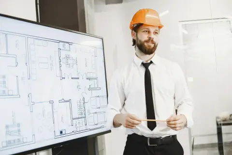 Mężczyzna w kasku pomarańczowym prezentuje projekt domu na ekranie. To szkolenie BHP dla pracownika, który zapoznaje się z planem.
