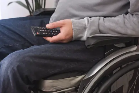Osoba na wózku inwalidzkim, z pilotem w dłoni, doświadcza umiarkowany stopień ograniczenia ruchomości, ale nadal cieszy się rozrywką.