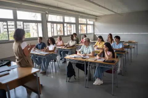 Nauczycielka przemawia do uczniów siedzących w ławkach w sali lekcyjnej.