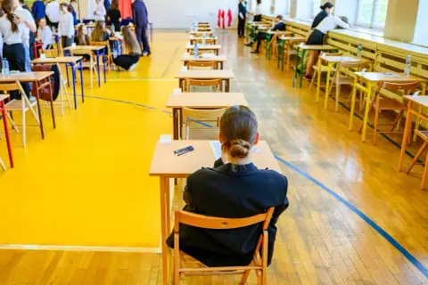 Uczniowie siedzą przy ławkach w sali gimnastycznej, przygotowując się do egzaminu dojrzałości.