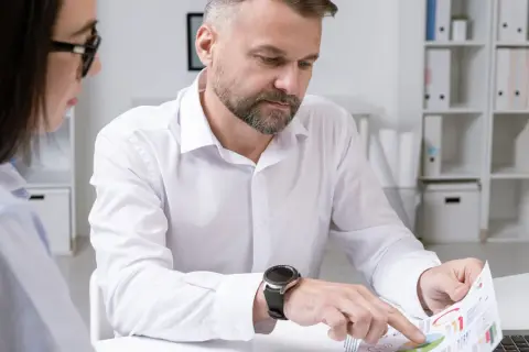 Mężczyzna z zarostem i zegarkiem na ręku analizuje wykresy, podczas gdy kobieta w okularach obserwuje. Ich praca wymaga ścisłego nadzoru.