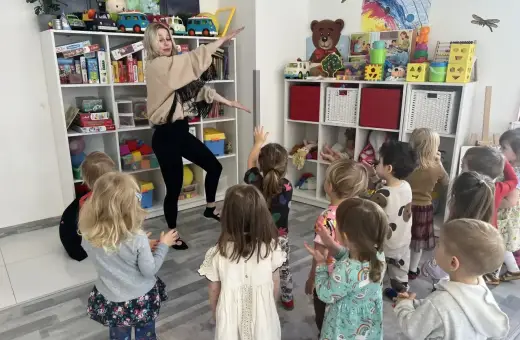 Nauczycielka prowadzi zajęcia z rytmiki w przedszkolu. Dzieci z uwagą obserwują jej ruchy, bawiąc się wesoło.
