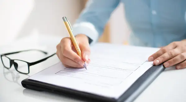 Dłoń pisze złotym długopisem na dokumencie, który wygląda jak aneks do umowy o pracę. Obok leżą okulary.