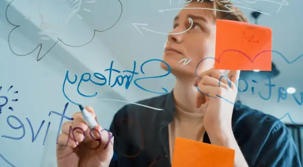 Młody mężczyzna pisze na szkle, tworząc schemat. Widać żarówkę symbolizującą pomysł i słowo "hit".