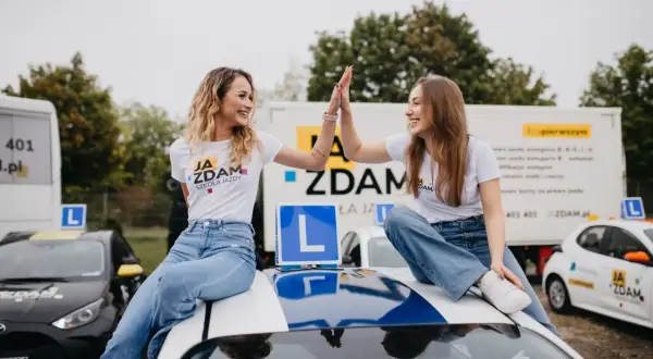 Dwie uśmiechnięte dziewczyny w koszulkach "Ja Zdam Szkoła Jazdy" świętują sukces, siedząc na dachu samochodu. To znak, że zdały egzamin klasyfikacyjny!