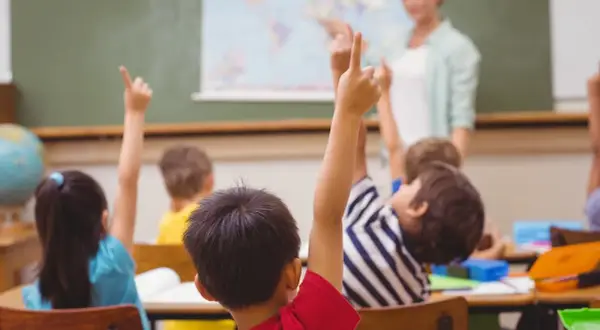 Uczniowie z podniesionymi rękami, gotowi do odpowiedzi. Nauczycielka ocenia ich zaangażowanie, co wpłynie na oceny z zachowania.