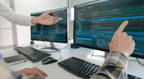 Dwóch programistów analizuje kod na monitorach. Jeden wskazuje palcem na ekran, drugi gestykuluje.