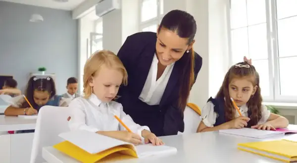Nauczycielka podczas hospitacji wspiera ucznia w pisaniu. Dzieci siedzą przy ławkach, skupione na zadaniu.