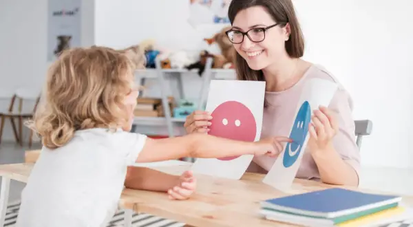 Uśmiechnięta psycholog z okularami pokazuje dziecku karty z buźkami. To część przygotowania pedagogicznego dla psychologów.