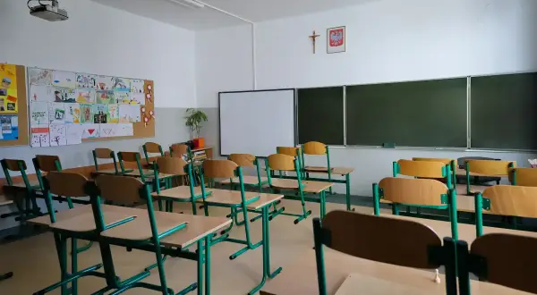 Pusta klasa szkolna z rzędami ławek i krzeseł. Na ścianie tablica, projektor i dziecięce rysunki. Cisza i spokój, jakby odwołane zajęcia w szkołach.