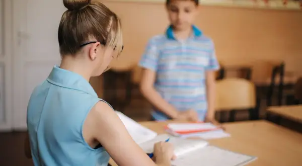 Nauczycielka z kucykiem przygląda się zeszytom, podczas gdy uczeń stoi w tle. Czy to zapowiedź podwyżki dla nauczycieli? Najnowsze wiadomości wkrótce.