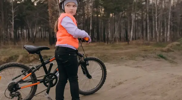 Dziewczynka w kasku i kamizelce przygotowuje się do egzaminu na kartę rowerową, stojąc obok swojego roweru na leśnej ścieżce.