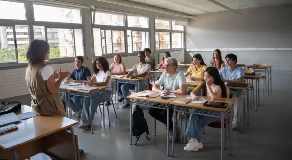 Nauczycielka prowadzi lekcję, a uczniowie siedzą w ławkach, słuchając jej uważnie. Wszyscy są zaangażowani w proces nauczania, a atmosfera jest pełna skupienia.