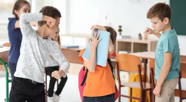 Chłopiec zasłania się zeszytem, podczas gdy inni uczniowie w klasie wydają się być zaangażowani w bullying.