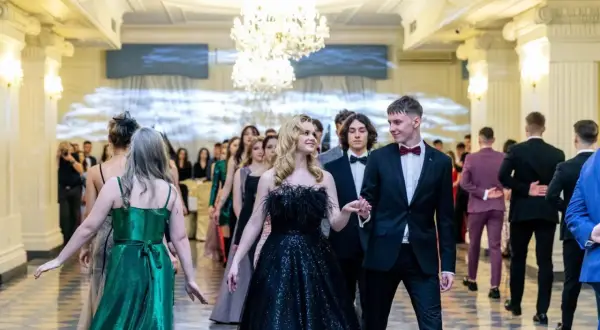 Młoda para idzie na studniówkę. Dziewczyna w czarnej, błyszczącej sukni z piórami, chłopak w eleganckim garniturze. W tle inni uczniowie w balowych strojach.