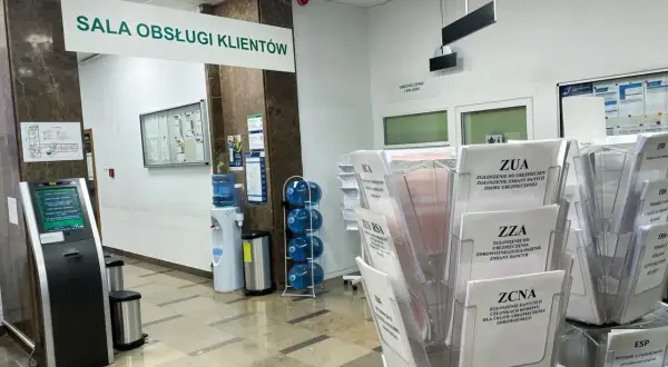 W sali obsługi klienta ZUS, obok dystrybutora wody, stoją stojaki z formularzami, m.in. ZUA, ZZA, ZCNA, RCA, ZUS RSA.