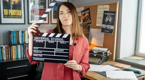 Kobieta trzyma klaps filmowy, gotowa do rozpoczęcia zajęć. W tle plakaty filmowe i maszyna do pisania, tworząc atmosferę pracy nad scenariuszem.