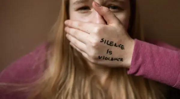 Przerażona dziewczyna zakrywa usta dłonią z napisem "Silence is violence". To obraz patologii społecznej, która odbiera głos.