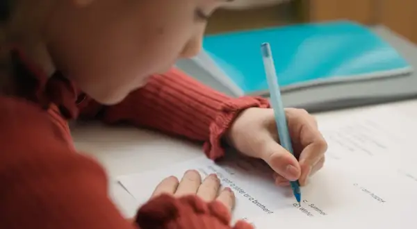 Dziecko w czerwonym swetrze pisze długopisem po karcie pracy, starając się o dopuszczający ocena.