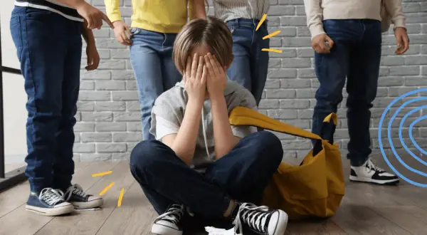 Chłopiec siedzi na podłodze, zakrywając twarz dłońmi, podczas gdy grupa dzieci stoi nad nim. To obraz tego, czym jest **bullying**.
