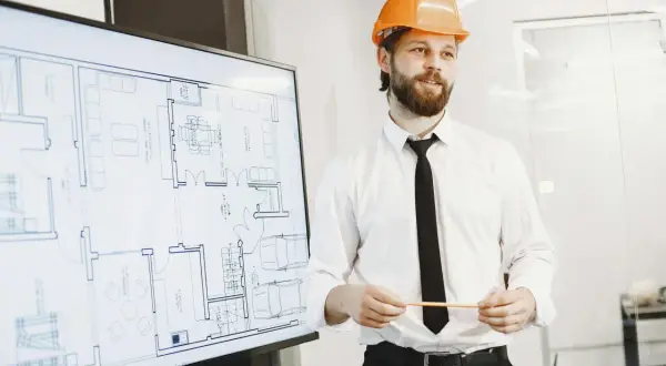Mężczyzna w kasku pomarańczowym prezentuje projekt domu na ekranie. To szkolenie BHP dla pracownika, który zapoznaje się z planem.