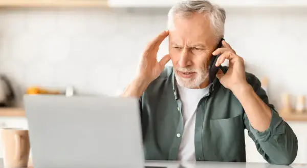 Zmartwiony mężczyzna rozmawia przez telefon, trzymając się za głowę. Obok laptopa i kubka. Wygląda na to, że jego urlop z powodu siły wyższej został zakłócony.