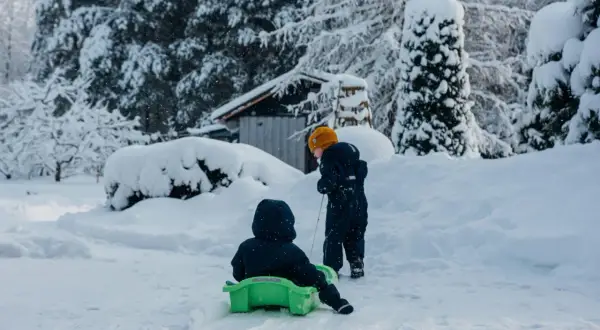 Dzieci cieszą się śnieżnymi feriami małopolskimi. Jedno zjeżdża na sankach, drugie ciągnie je przez zasypany śniegiem ogród.