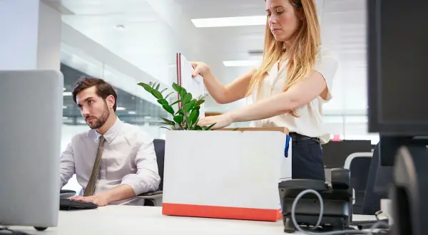 Kobieta pakuje rzeczy do kartonu, co sugeruje likwidację stanowiska pracy. Obok siedzi zamyślony mężczyzna przy komputerze.